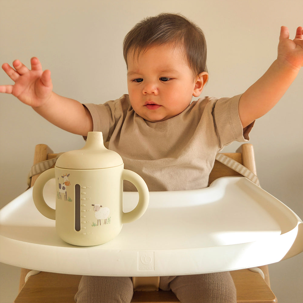 Child in a high chair with a beige cup on a tray, against a neutral background 