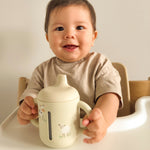 Baby sitting in a high chair holding a white cup with a lid. 