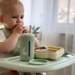 Child sitting at a high chair with a green cup and utensils on a tray 
