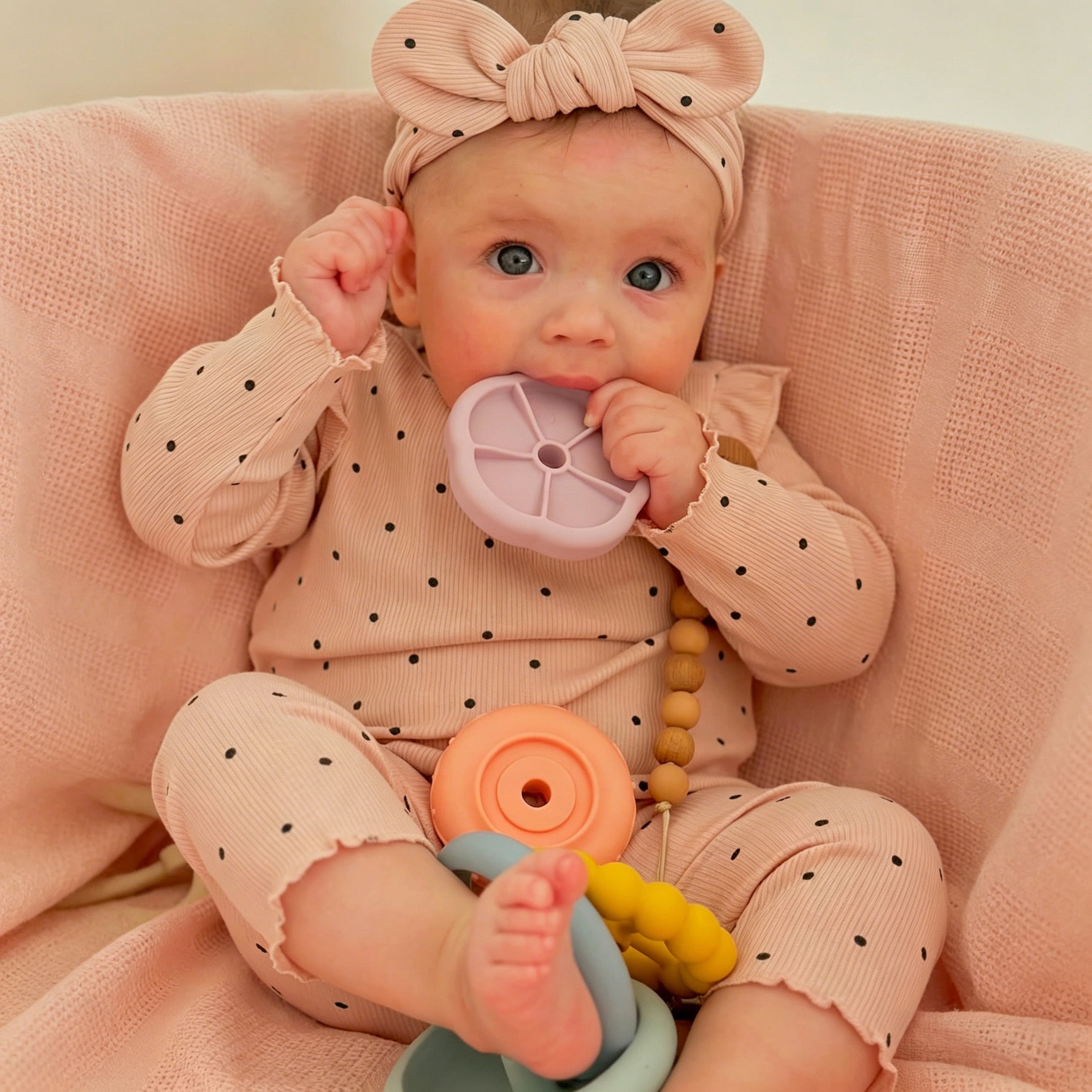 Baby in a polka dot outfit with toys on a pink blanket 