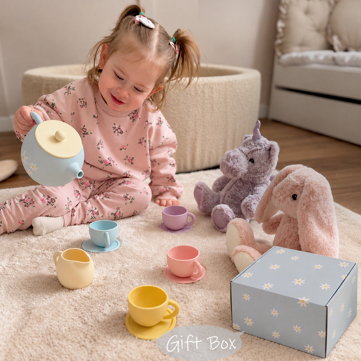 A toddler in pink floral pajamas sits on a cream rug playing with a pastel tea party set, including a light blue teapot, cups, a yellow mug, and a matching gift box. A purple plush unicorn and a pink plush bunny are beside her in a softly lit room.