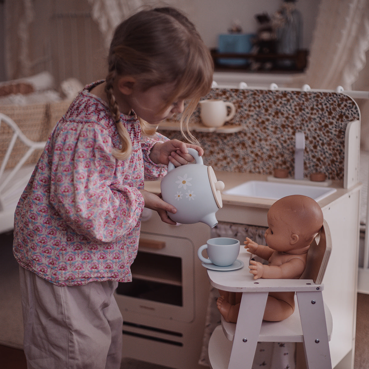 A young girl in a pink floral long-sleeve shirt and beige pants plays with a doll in a toy kitchen, pouring tea from a light blue teapot into a cup on a saucer held by the doll seated in a white high chair.