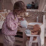 A young girl in a pink floral long-sleeve shirt and beige pants plays with a doll in a toy kitchen, pouring tea from a light blue teapot into a cup on a saucer held by the doll seated in a white high chair.