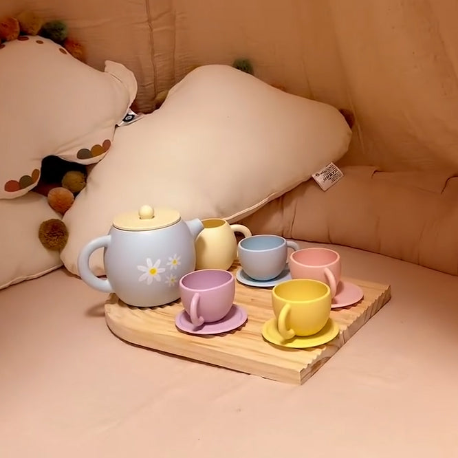 Set of colorful ceramic tea cups and a teapot on a wooden tray with pillows in the background.