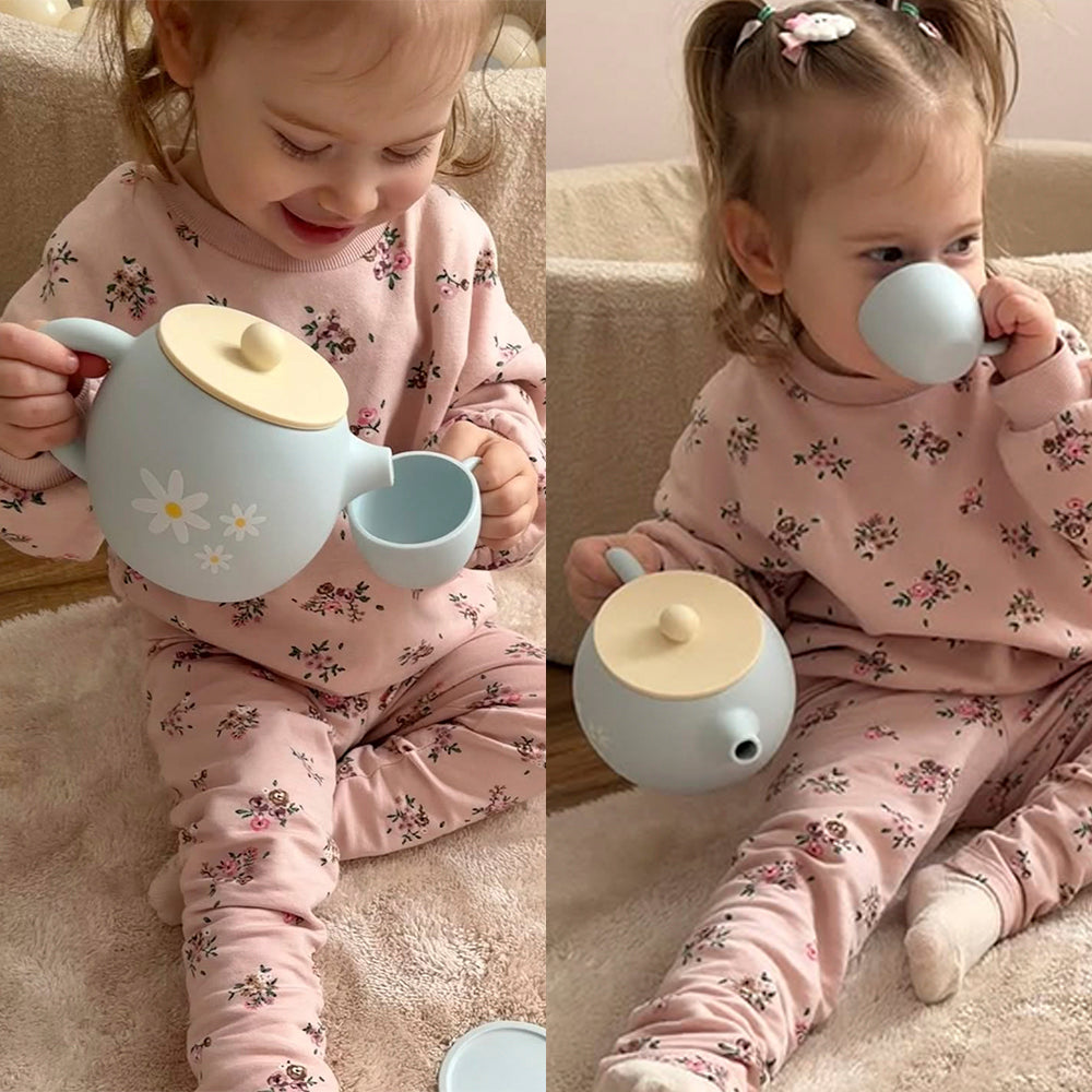 Child in pink floral pajamas playing with a toy teapot and cup on a carpeted floor.